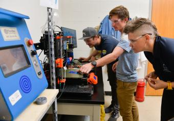 Two students with professor look at 3d printer