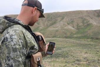Man in camo looks at phone with foothills in distance