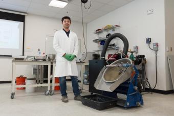 A researcher in a white lab coat and green gloves stands next to specialized equipment used for mine tailings research inside a well-equipped laboratory.