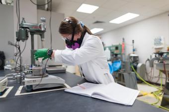 A student wearing safety goggles, gloves, and a respirator mask performs a lab experiment in a mineral processing laboratory, measuring material on a scale next to an open lab notebook.
