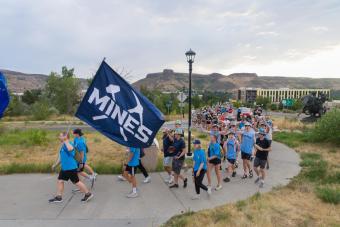 crowd of students walk on sidewalk with campus in background and Mines flag in foreground