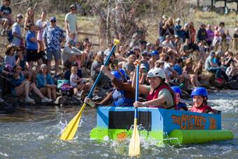 students in helmets paddle Mystery Machine-themed boat made of cardboard in creek with spectators on banks