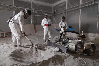 three students in white protective suits stand on gray powdery material in lunar surface simulator