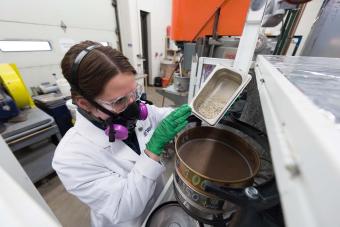 female researcher in respirator and white lab coat pours rocky material from one container to larger one