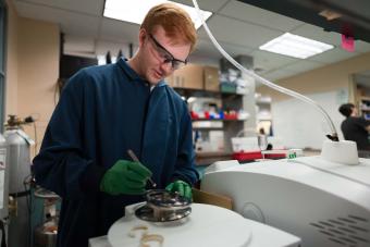 male student wearing safety glasses and blue lab coat works in the lab