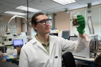 aaron goodman in a white lab coat holding a glass vial containing brown minerals