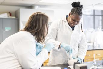 two female students in white lab coats work in lab