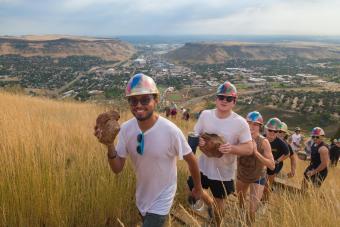line of students wearing hardhats and holding rocks walk up a grassy hill during M Climb