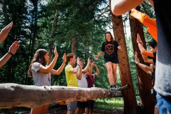 student walks across a log balance beam with fellow students standing on either side with hands up