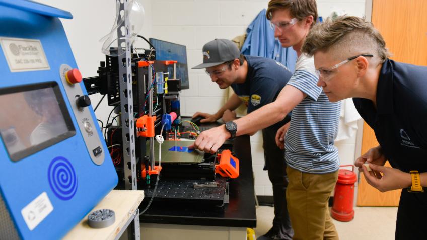Two students with professor look at 3d printer