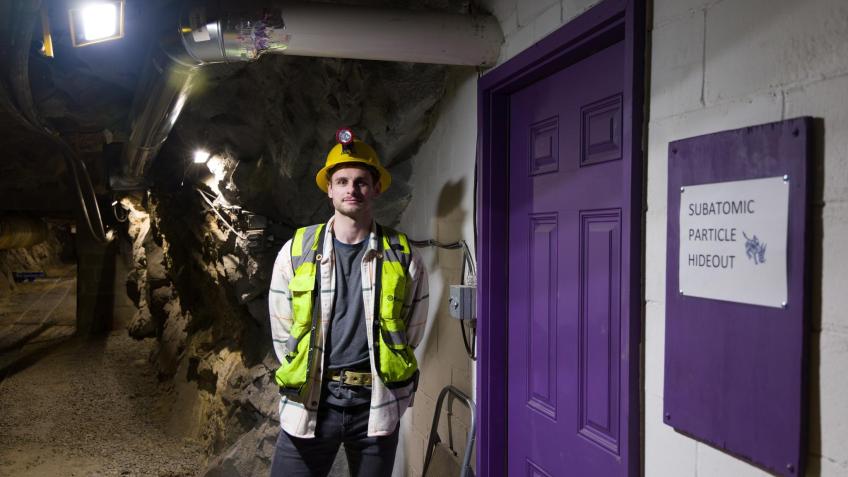 Male student in safety gear poses by purple door inside underground mine