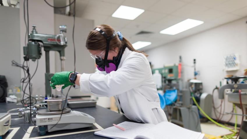 A student wearing safety goggles, gloves, and a respirator mask performs a lab experiment in a mineral processing laboratory, measuring material on a scale next to an open lab notebook.