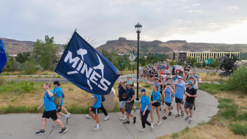 crowd of students walk on sidewalk with campus in background and Mines flag in foreground