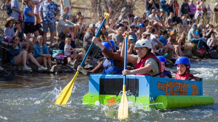 students in helmets paddle Mystery Machine-themed boat made of cardboard in creek with spectators on banks