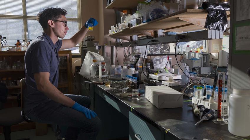 male researcher sitting at lab bench in front of window looks at liquid in vial