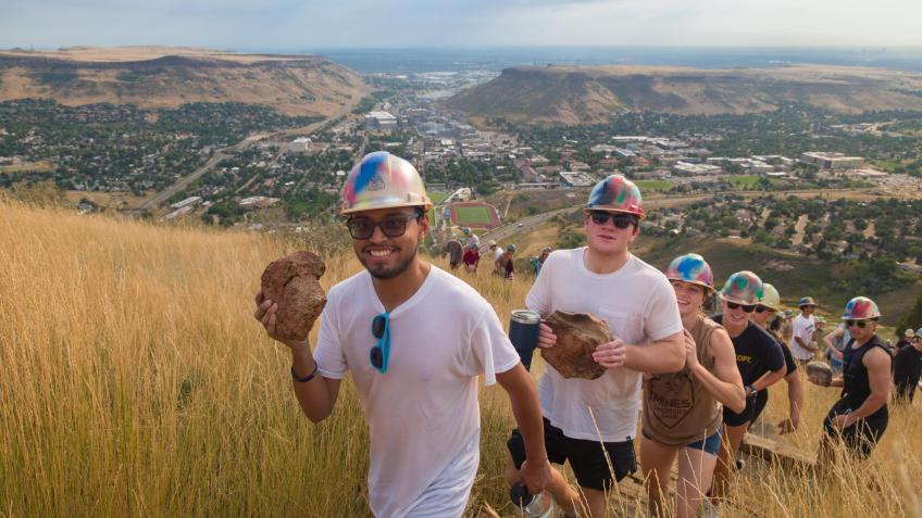 line of students wearing hardhats and holding rocks walk up a grassy hill during M Climb