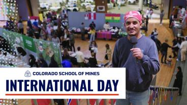 A student holds a microphone and smiles at the camera at Colorado School of Mines’ International Day event. In the background, a large room is filled with booths displaying international flags and food tables, with students and community members gathered to sample dishes and explore cultural exhibits. A banner at the bottom reads "Colorado School of Mines International Day.