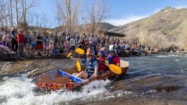 students in cardboard boat floating down creek with mount zion in background