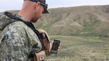 Man in camo looks at phone with foothills in distance