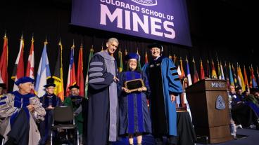 female PhD graduate in regalia holds award between President Johnson and Vice President of Research Walt Copan