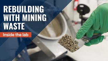A gloved hand holds a small metal scoop of processed mine tailings in a laboratory setting. Behind it, lab equipment and a mixing drum are visible. Overlaid text reads: “Rebuilding with Mining Waste – Inside the lab.”
