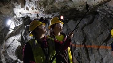 Two Colorado School of Mines students wearing safety vests, hard hats, and headlamps examine the rock ceiling inside the Edgar Experimental Mine using a mounted inspection camera. The underground setting is lit with work lights, revealing rough stone walls.