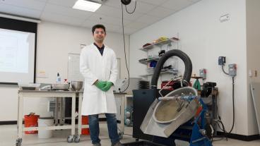 A researcher in a white lab coat and green gloves stands next to specialized equipment used for mine tailings research inside a well-equipped laboratory.