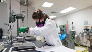 A student wearing safety goggles, gloves, and a respirator mask performs a lab experiment in a mineral processing laboratory, measuring material on a scale next to an open lab notebook.