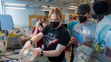 four students wearing masks pour water in concrete mix in lab