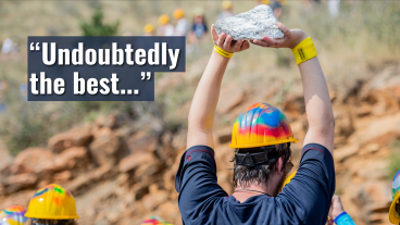 Student in a hard hat holds a rock overhead during the M Climb, with on-screen text that reads, “Undoubtedly the best…”