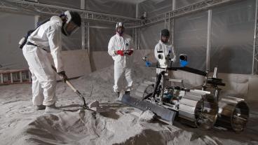 three students in white protective suits stand on gray powdery material in lunar surface simulator