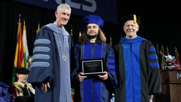 three men in graduation regalia pose together with plaque in the center