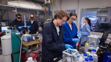 four students in lab coats work at various stations in a lab