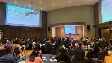 large room full of people sitting at tables with math problem projected on screen