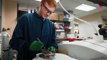 male student wearing safety glasses and blue lab coat works in the lab