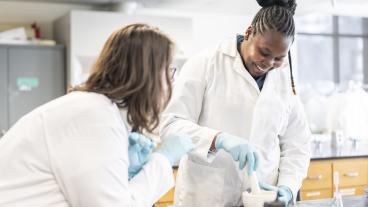 two female students in white lab coats work in lab