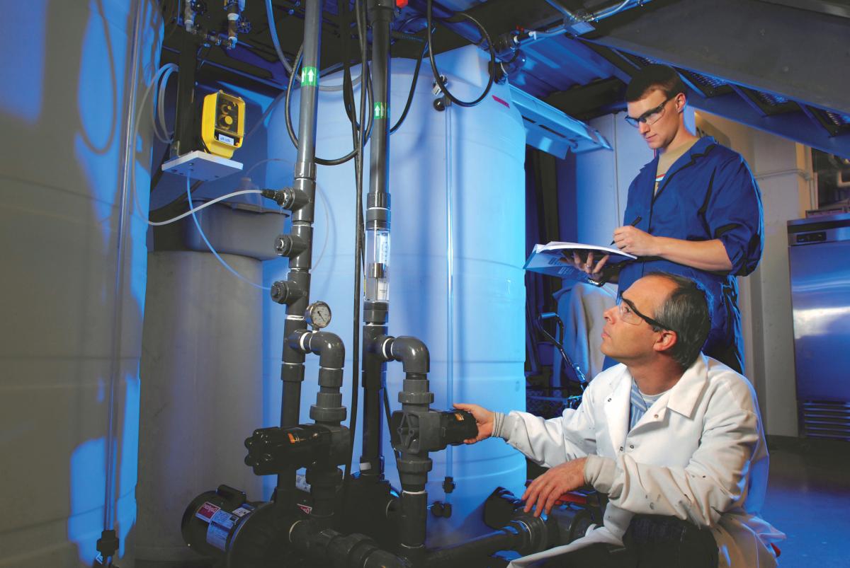 Water pipes with electronic gauges are shown, as the professor kneels to read the information and a student records the data.